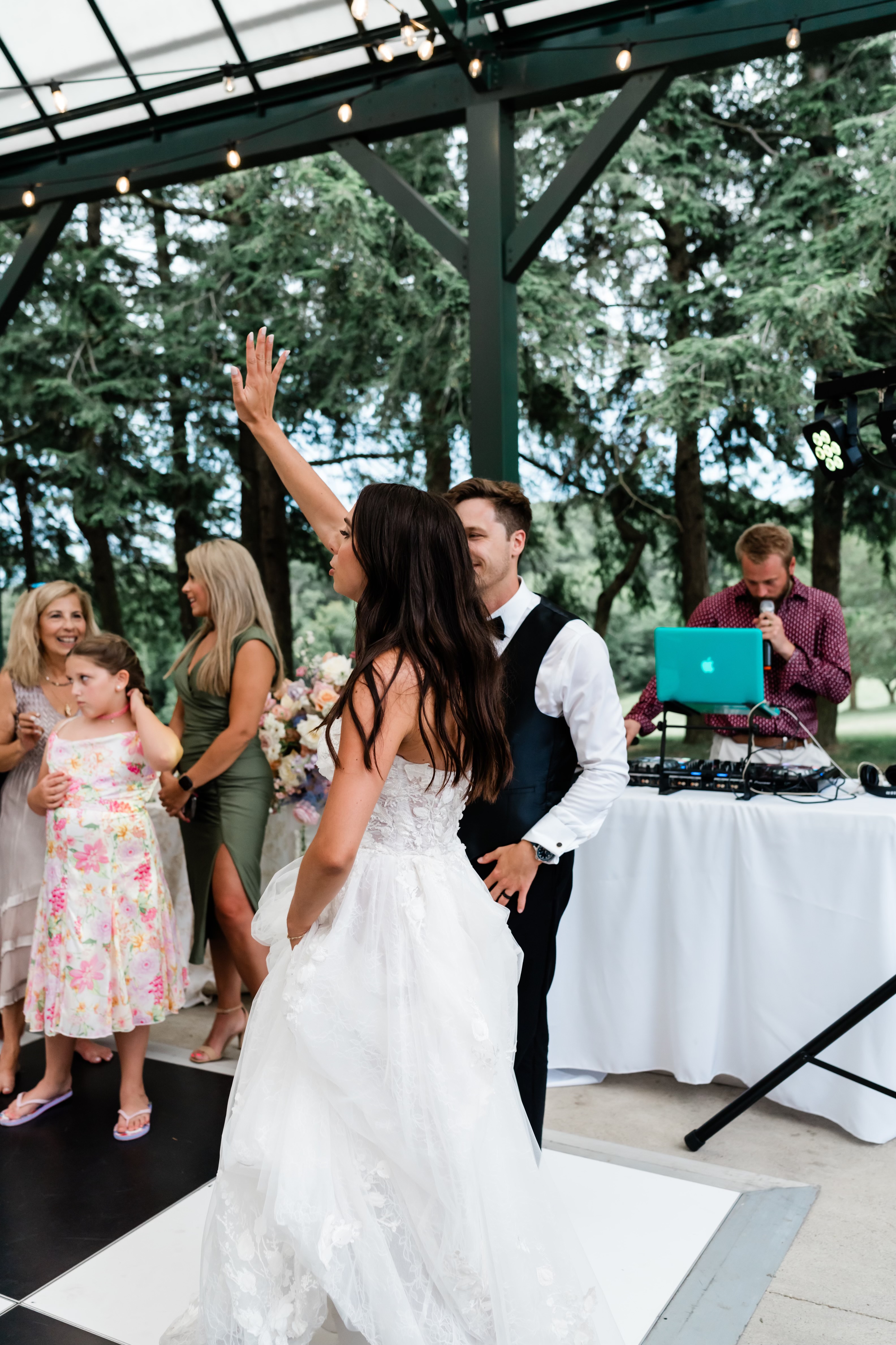 Bride and groom dancing in the foreground while the DJ works at the booth behind, glass pavilion venue in daylight.