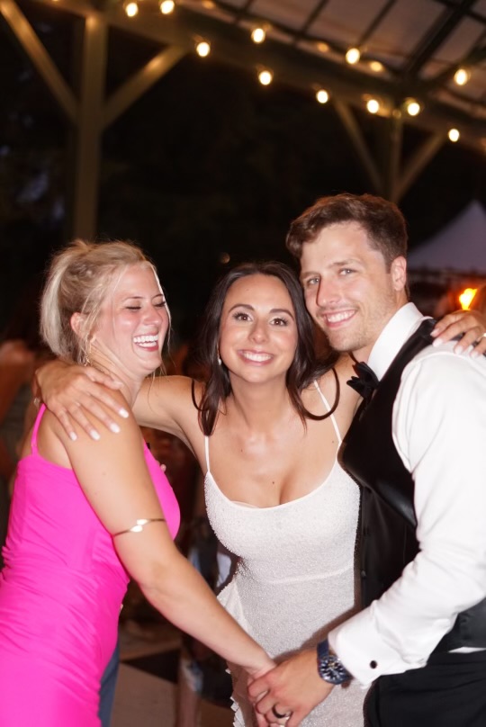 Bride, groom, and laughing maid of honor in close-up at an outdoor evening reception under string lights.