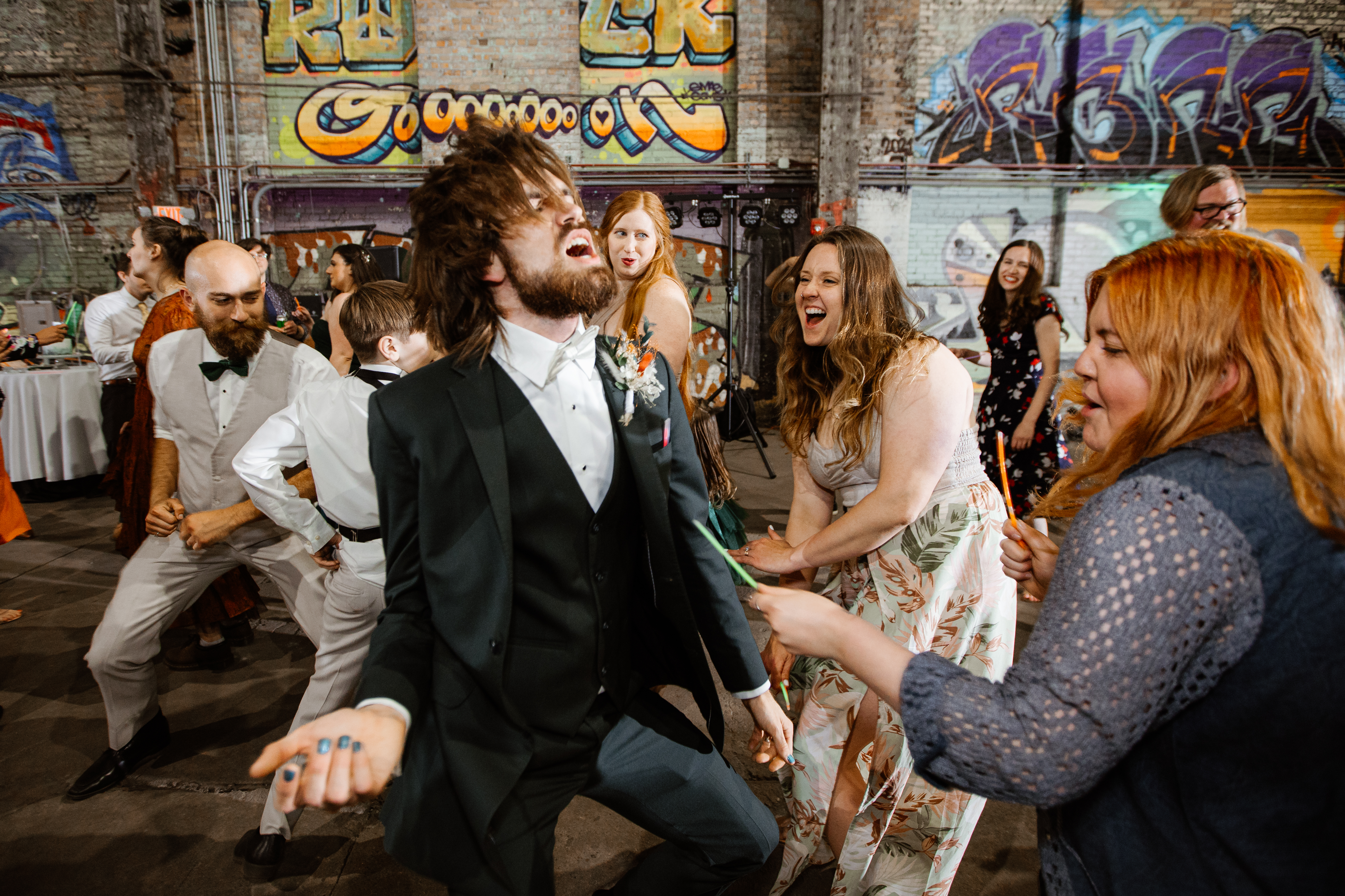 High-energy candid of groom mid-dance at an industrial venue with graffiti walls, crowd around him cheering.