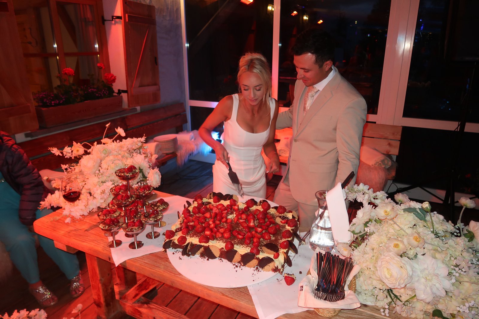Bride and groom cutting a strawberry-topped cake together at an Italian wedding reception, string lights and rustic wood shutters behind them.
