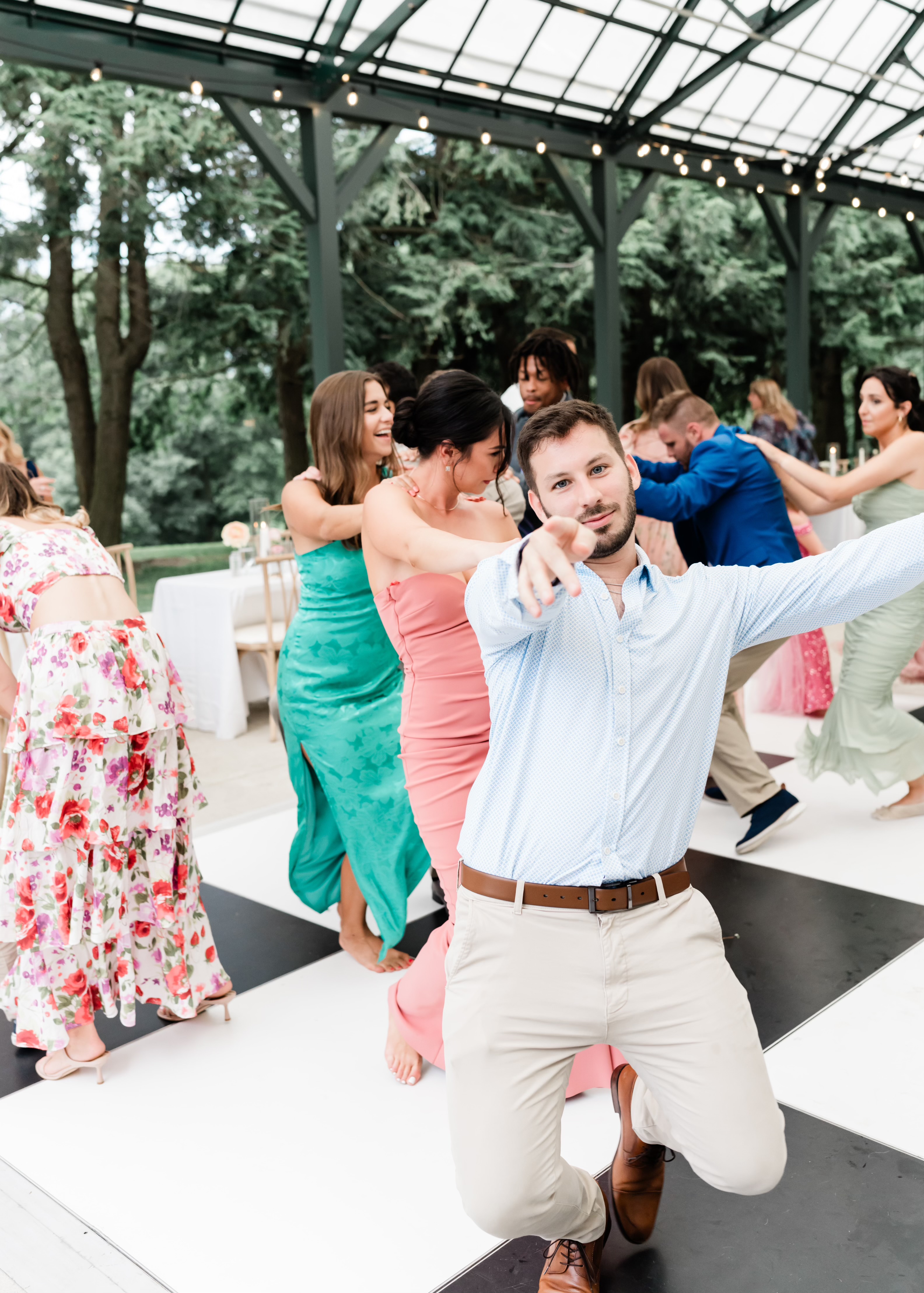 Guest mid-dance pointing at camera, other guests dancing in colorful dresses behind, glass pavilion venue in daylight.