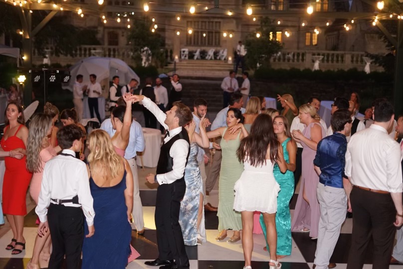 Full wedding reception dance floor under string lights at an outdoor estate venue, mansion in the background at dusk.