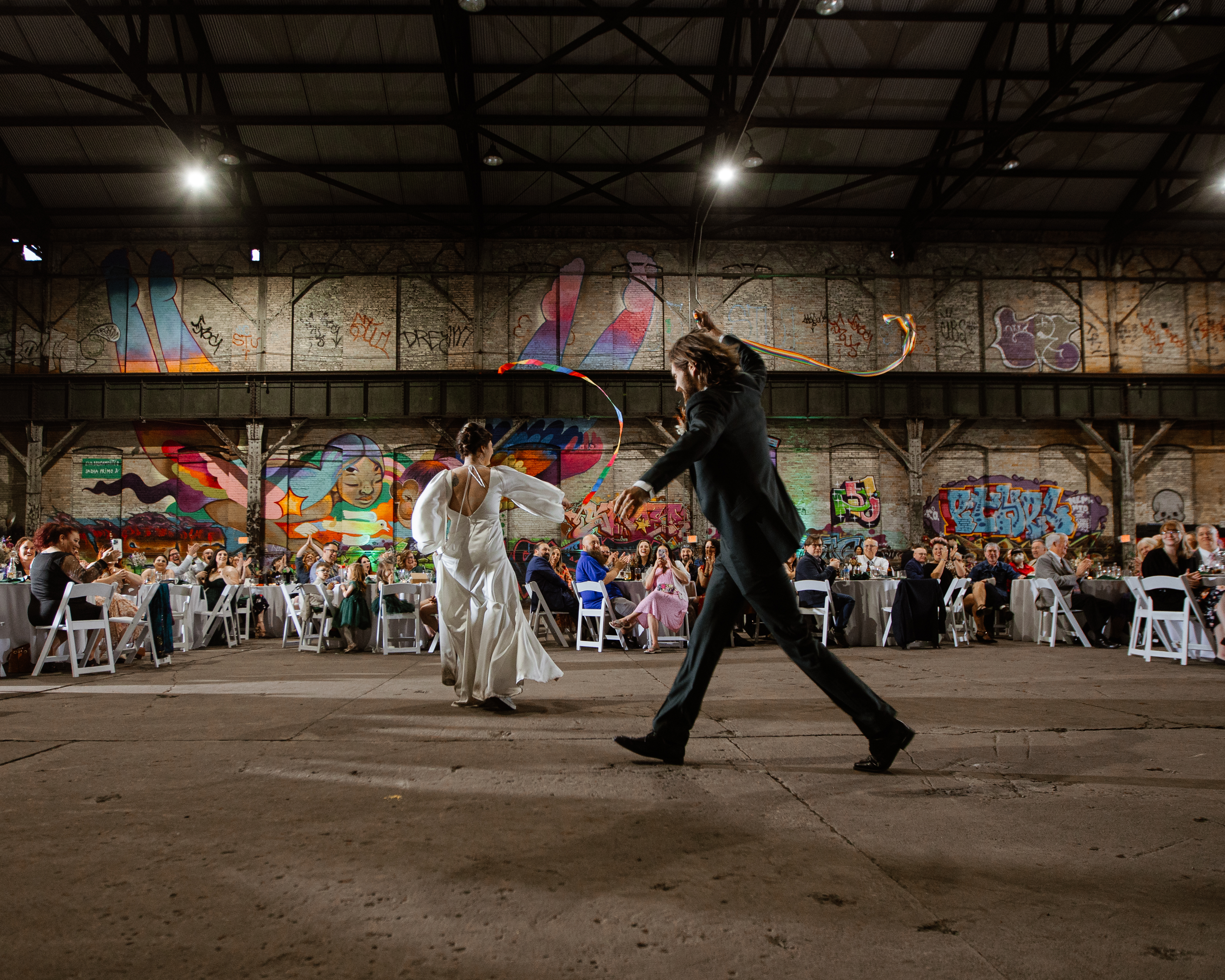 Couple dancing with flowing ribbons at an industrial venue, guests seated around them watching.