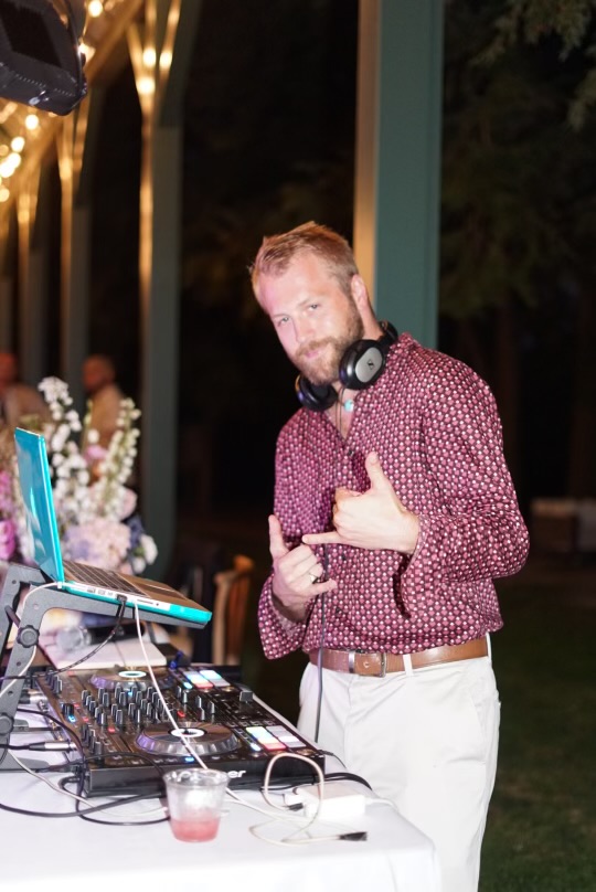 DJ Willionaire at the mixing booth during a wedding reception, headphones around his neck, soft warm evening light.